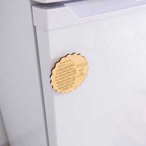 May include: A wooden magnet with a decorative scalloped edge. The magnet features a gold-colored background with Arabic text and the words "Ayatul Kursi" in English. The magnet is attached to a white refrigerator.