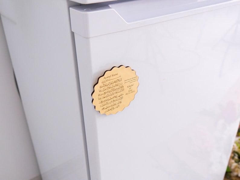 May include: A wooden magnet with a decorative scalloped edge. The magnet features a gold-colored background with Arabic text and the words "Ayatul Kursi" in English. The magnet is attached to a white refrigerator.