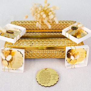May include: Gold decorative boxes with white lids, each containing a round, gold-plated plaque with Arabic calligraphy. The boxes are arranged on a gold-plated tray with a decorative pattern. The plaques are tied with white ribbon and decorated with dried flowers.