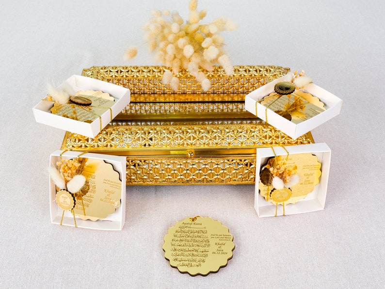 May include: Gold decorative boxes with white lids, each containing a round, gold-plated plaque with Arabic calligraphy. The boxes are arranged on a gold-plated tray with a decorative pattern. The plaques are tied with white ribbon and decorated with dried flowers.
