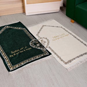 Könnte beinhalten: Zwei Gebetsteppiche, einer dunkelgrün und einer cremefarben, mit dekorativen Bordüren und Fransen. Auf dem grünen Teppich ist der Name "Ibrahim" eingestickt, auf dem cremefarbenen Teppich der Name "Fatima".