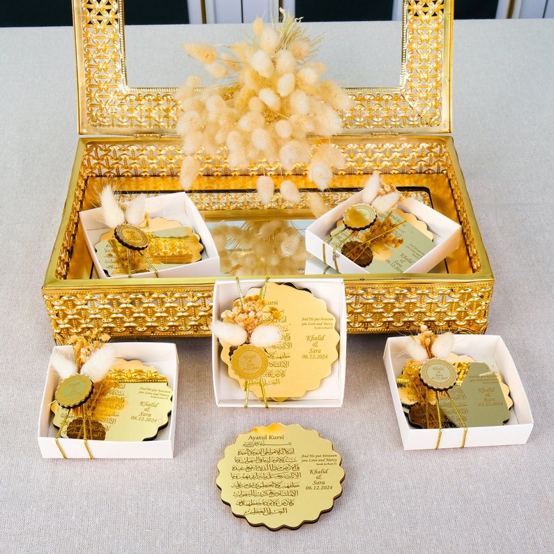 May include: A set of five gold-colored, decorative, round plaques with Arabic calligraphy and the names "Khalid & Sara" and the date "06.12.2023". The plaques are arranged in a gold-colored box with a white lining. The box is decorated with white flowers.