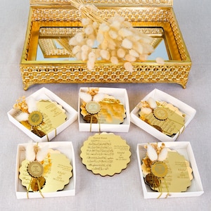 May include: Five gold-colored, round, decorative plaques with Arabic calligraphy and a gold seal. Each plaque is in a white box with a small bouquet of dried white flowers. The plaques are arranged in a row on a white surface.