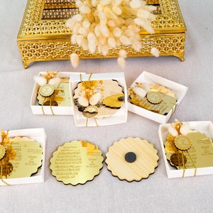 May include: Six white boxes with gold trim contain round, gold-colored, decorative plaques with Arabic calligraphy and a small, gold-colored seal. The plaques are in various stages of being opened, revealing the gold-colored backing. One box contains a plain, wooden plaque.