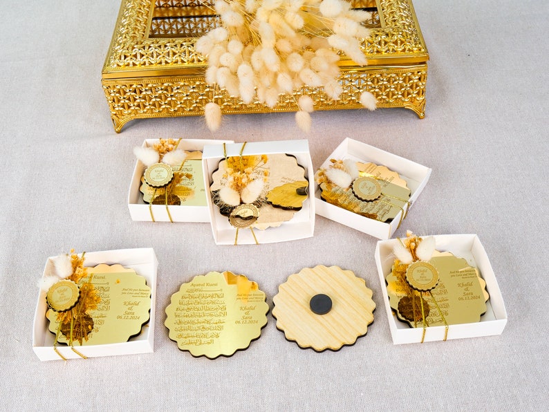 May include: Six white boxes with gold trim contain round, gold-colored, decorative plaques with Arabic calligraphy and a small, gold-colored seal. The plaques are in various stages of being opened, revealing the gold-colored backing. One box contains a plain, wooden plaque.