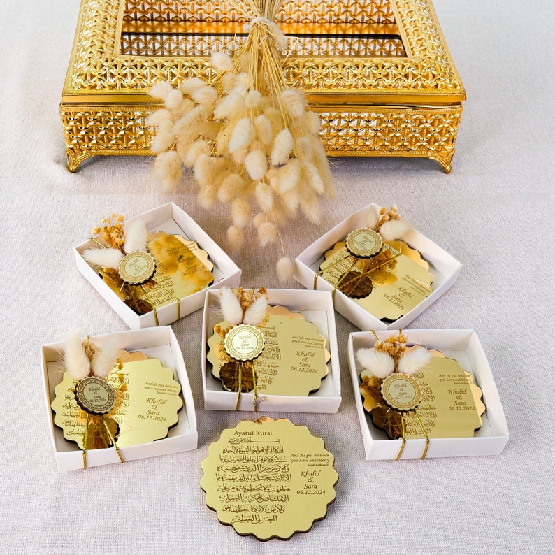 May include: Gold-colored decorative plaques with Arabic calligraphy and a gold seal. The plaques are in white boxes with dried flowers. The text on the plaques reads "Khalid & Sara 06.12.2024".