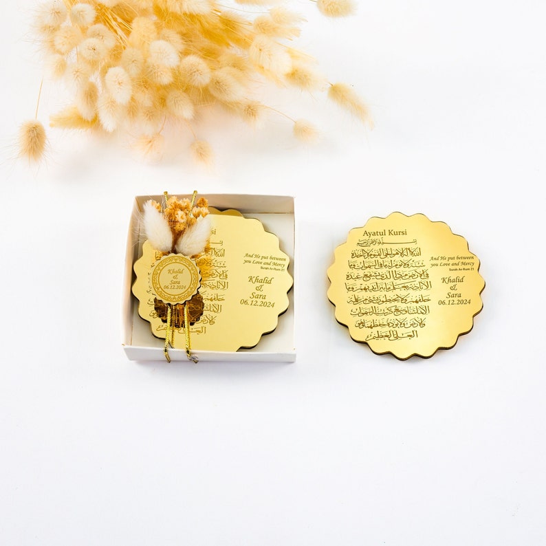 May include: Two gold-colored round wooden plaques with Arabic calligraphy and the text "Ayatul Kursi" and "And He put between you Love and Mercy". The plaques are in a white box with a small bouquet of dried flowers.