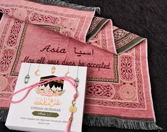 May include: A pink prayer rug with ornate patterns and the word "Asia" in English and Arabic script. A white box with a pink beaded prayer necklace and the words "Umrah Mubarak" is also present. The text "May all your duas be accepted" is written on the rug.