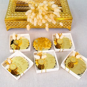May include: Six white boxes with gold trim, each containing a round gold mirror with Arabic calligraphy and a small bouquet of dried white flowers. The boxes are arranged in two rows of three.