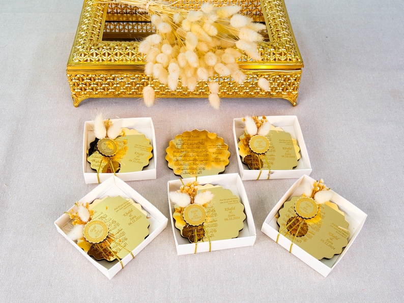 May include: Six white boxes with gold trim, each containing a round gold mirror with Arabic calligraphy and a small bouquet of dried white flowers. The boxes are arranged in two rows of three.
