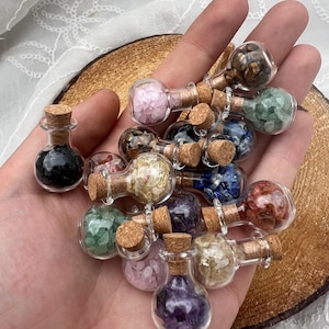 May include: A selection of miniature glass bottles, each filled with different coloured gemstones and sealed with a cork. The bottles contain pink, green, purple, and brown stones. The bottles are held in a hand.
