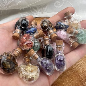 May include: A selection of miniature glass bottles with corks, filled with various coloured gemstones. The bottles are in different shapes, including heart-shaped, and the gemstones are in shades of purple, pink, yellow, and blue.