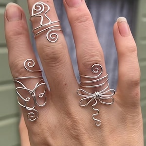 May include: A hand displays four silver wire rings. The rings feature unique designs, including a butterfly, a dragonfly, and abstract shapes. The rings are crafted from thin, coiled wire, showcasing a delicate and artistic aesthetic.