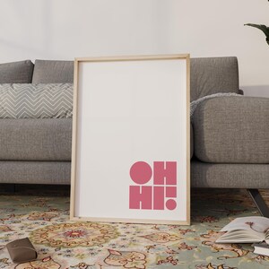 May include: A framed art print with a light wood frame. The print features the words "OH HI!" in pink, geometric shapes on a white background. The artwork is propped against a grey sofa on a patterned rug.