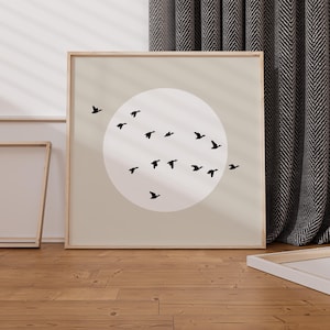 May include: A framed artwork featuring a beige background with a large white circle. Silhouetted black birds fly in a curved formation across the circle. The frame is a light wood color, and the artwork is displayed on a wooden floor.