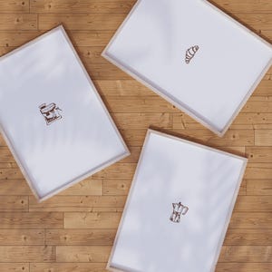 May include: Three framed prints with light wood frames on a wooden surface. Each print features a brown line drawing: a coffee maker, a croissant, and a coffee grinder. The prints are arranged at different angles.