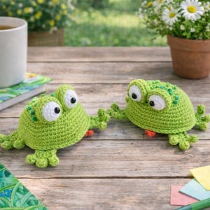 May include: Two handmade green crochet frog toys with large white eyes and orange accents are displayed on a wooden table. The frogs are positioned near a cup of tea, a notebook, and a pot of daisies, suggesting a whimsical theme.