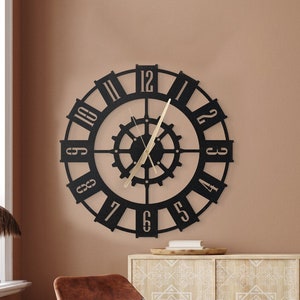 Könnte beinhalten: Eine schwarze Metalluhr mit römischen Ziffern und einem Zahnrad-Design hängt an einer braunen Wand. Zwei braune Ledersessel und ein brauner Lederhocker stehen vor einem hellen Holzkasten mit einem weißen geometrischen Muster.