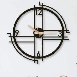 Peut inclure: Une horloge murale moderne en métal noir avec un design circulaire et des chiffres minimalistes à 3, 6, 9 et 12. Les aiguilles de l'horloge sont de couleur bois clair. L'horloge est fixée sur un mur blanc au-dessus d'un meuble en bois.