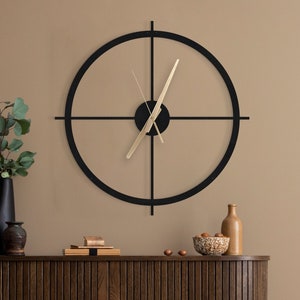 Op de afbeelding: Een zwarte en gouden klok met een ronde lijst en vier spaken. De klok hangt aan een beige muur boven een donkerhouten kast.