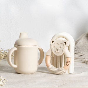 May include: A beige silicone sippy cup with two handles and a lid, next to a beige silicone teether with a wooden ring and a bear face design. The teether is on a beige silicone stand.