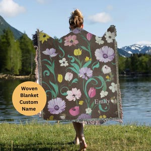 May include: A woven blanket with a floral pattern in shades of pink, purple, and green. The blanket has a brown background and the text "Emily" is woven into the design. The blanket is being held by a person.