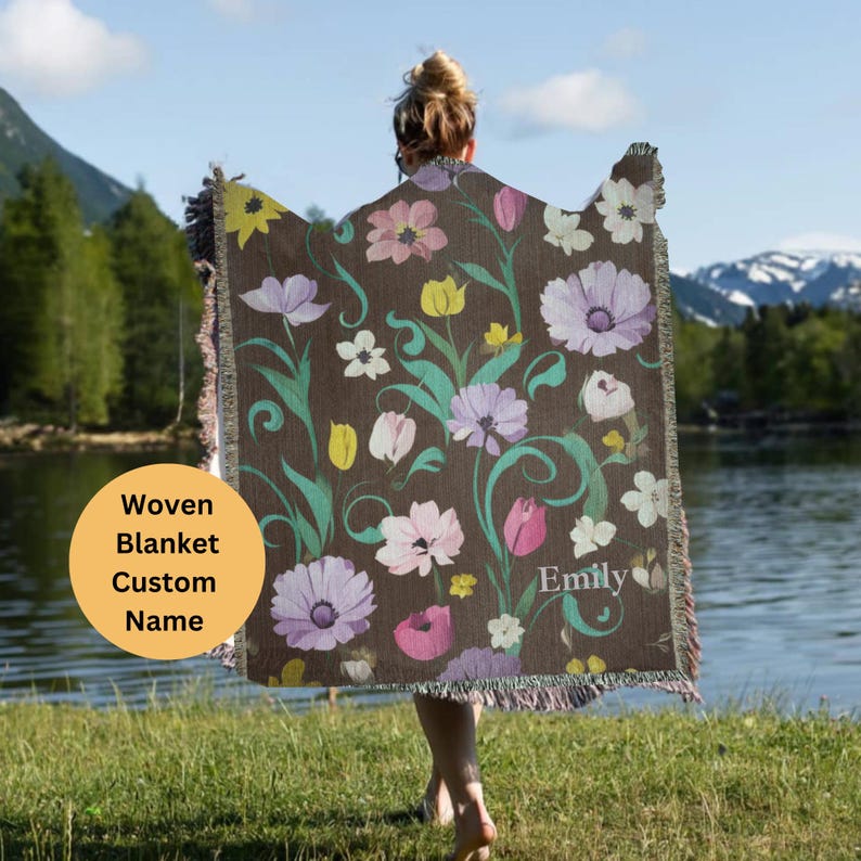 May include: A woven blanket with a floral pattern in shades of pink, purple, and green. The blanket has a brown background and the text "Emily" is woven into the design. The blanket is being held by a person.