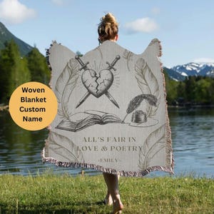 May include: A woven blanket with a gray background featuring a broken heart with two swords crossing through it, a book, a quill pen, and an inkwell. The text "All's fair in love & poetry -EMILY- " is printed below the image.