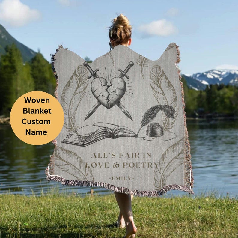 May include: A woven blanket with a gray background featuring a broken heart with two swords crossing through it, a book, a quill pen, and an inkwell. The text "All's fair in love & poetry -EMILY- " is printed below the image.