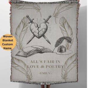 May include: A woven throw blanket with a gray background and a black and white illustration of a broken heart, two swords, a book, a quill pen, and an inkwell. The text "All's Fair in Love & Poetry -Emily- " is printed in gold on the blanket.