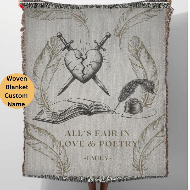 May include: A woven throw blanket with a gray background and a black and white illustration of a broken heart, two swords, a book, a quill pen, and an inkwell. The text "All's Fair in Love & Poetry -Emily- " is printed in gold on the blanket.