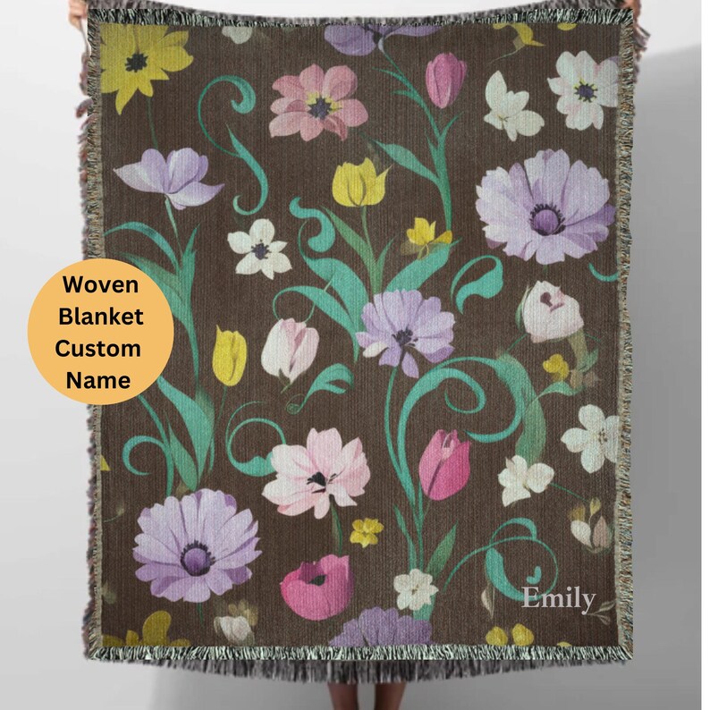 May include: A woven blanket with a floral pattern in shades of pink, purple, yellow, and green on a brown background. The blanket has a fringe border and the name "Emily" is woven into the bottom right corner.