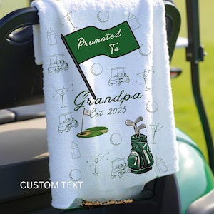 Op de afbeelding: Witte golfhanddoek met groene accenten en de tekst "Promoted to Grandpa Est 2025". De handdoek heeft golf-thema illustraties, waaronder golfkarren, golfballen en een golftas. Een groene vlag leest "Promoted To".