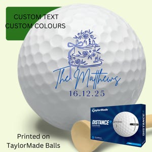 May include: White golf ball with a blue floral cake design, the text "The Matthews" and the date 16.12.25. The ball is printed on a TaylorMade ball. A box of TaylorMade golf balls is also visible.