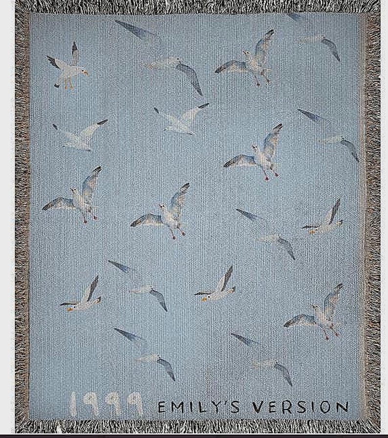 May include: A blue woven tapestry with a white fringe border. The tapestry features a repeating pattern of white seagulls flying against a light blue background. The text "1999 EMILY'S VERSION" is printed in black at the bottom of the tapestry.