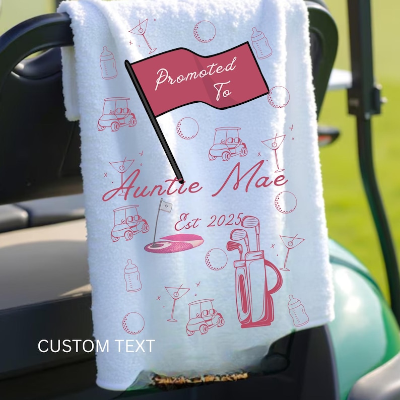 Op de afbeelding: Witte golfhanddoek met roze illustraties van golfkarren, golfballen en martiniglazen. De handdoek heeft de tekst "Promoted To Auntie Mae Est 2025". De woorden "Custom Text" staan onderaan.