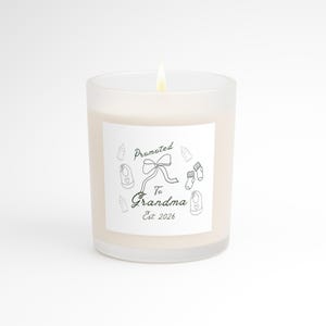 Op de afbeelding: Een brandende, witte kaars in een matglazen pot. De kaars heeft een wit etiket met de tekst "Promoted To Grandma Est 2026" en baby-thema illustraties. De kaars staat op een witte achtergrond.
