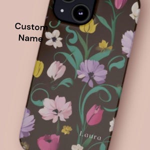 May include: A brown phone case with a floral pattern featuring pink, purple, and yellow flowers. The case has a cutout for the camera lens and the text "Laura" is printed on the bottom.
