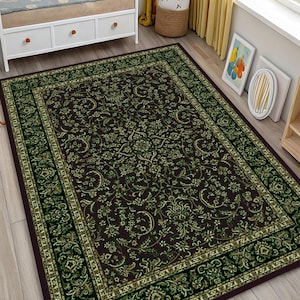 Puede incluir: Una alfombra rectangular con un centro marrón oscuro y un estampado floral verde y dorado. La alfombra tiene un borde verde oscuro con un diseño repetitivo. La alfombra está colocada sobre un suelo de madera clara.