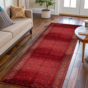 May include: A long, rectangular red rug with an intricate geometric pattern. The rug features a central field of repeating squares and rectangles, bordered by a detailed design. The rug is placed on a wooden floor, adding warmth and colour to the space.