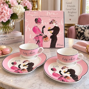 Puede incluir: Un juego de té para dos con dos tazas de cerámica rosa y blanca y platos a juego con bordes festoneados. Cada pieza está decorada con ilustraciones de dos figuras y el texto "très chic". El set viene en una caja rosa "Ladies Who Lunch".