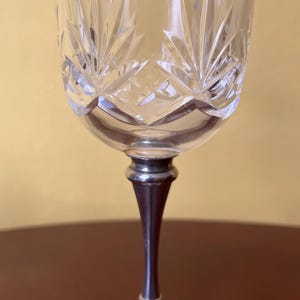 May include: A clear crystal wine glass with a decorative etched pattern. The glass has a silver stem and base. The etched design features a series of pointed shapes radiating from the center of the bowl. The glass is designed for serving wine.
