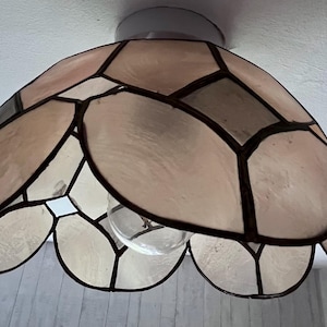 May include: A ceiling light fixture with a stained glass lampshade. The lampshade is composed of petal-shaped panels in shades of pink, beige, and gray, outlined with dark brown lines. The light bulb is visible through the bottom of the shade.
