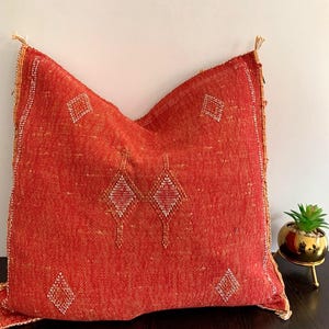 May include: A square, red decorative pillow with a diamond pattern. The pillow has a textured weave and a fringe trim. The diamond pattern is embroidered with white and gold thread. The pillow is approximately 20 inches square.