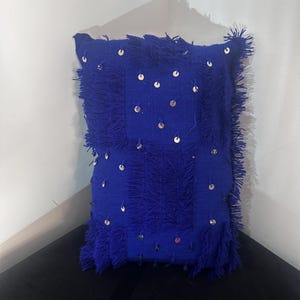 Puede incluir: Un cojín cuadrado azul real con flecos y lentejuelas plateadas dispersas. El cojín está hecho de una tela texturizada y está colocado en una esquina.