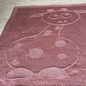 Könnte beinhalten: Eine gestrickte Decke in einem staubigen Rosaton mit einem Giraffenmuster. Die Giraffe hat große Augen, Ohren und Flecken. Die Decke hat ein strukturiertes Aussehen und einen detaillierten Rand.