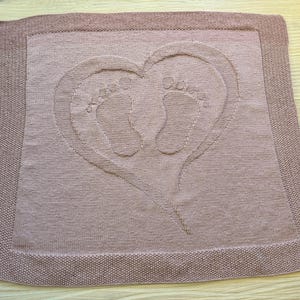 May include: A dusty rose-colored baby blanket with a heart-shaped design. The heart contains two baby footprints. The blanket has a textured border and is made of a soft material.