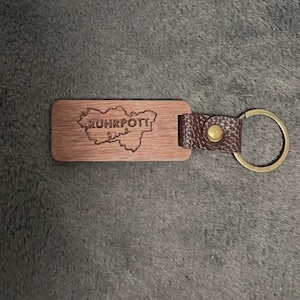 Könnte beinhalten: Ein Holzschlüsselanhänger mit einem braunen Lederband und einem Metallring. Der Holzanhänger zeigt eine Karte der Ruhrpott-Region mit dem eingravierten Text "Ruhrpott" und "ling".