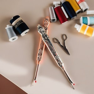 May include: Two ornate metal thread cutters with floral designs and the words "Embroidery" and "Hermès Paris" on the handles. The cutters are on a light beige surface with spools of thread and a pair of small scissors.