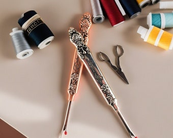 May include: Two ornate metal thread cutters with floral designs and the words "Embroidery" and "Hermès Paris" on the handles. The cutters are on a light beige surface with spools of thread and a pair of small scissors.